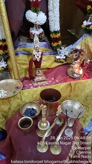 Photograph of a small shrine table during a ritual setup. Centre stands a small brass-coloured deity murti of a standing female figure wearing a red garment, a white flower necklace and a tall decorated crown, flanked by white and orange flower garlands hanging behind. Before it sit a copper vessel filled with liquid, a small brass bowl of dark blue crystals, a brass cup, a tall brass oil lamp stand, and a lit oil lamp on a brass plate to the right. Marigold and petals are scattered on the gold and red cloth. No people are visible.