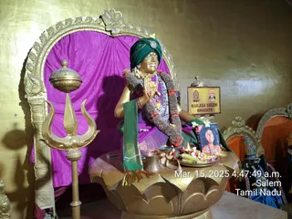 A golden statue of a seated male figure with hands joined in namaskar, wearing a green turban with a brooch, a garland of marigolds and orange flowers, and a magenta cloth; a golden trident stands to the left and a golden lotus-shaped pedestal holds a kalasha, oil lamps, a plate of flowers and fruit, and a framed photo of a smiling man. A golden arch frames a magenta backdrop. A sign reads KAILASA SALEM BHARATH. A timestamp in the corner shows Mar. 15, 2025 4:47:49 a.m., Salem, Tamil Nadu. The figure is a statue, and the framed photo is a printed picture, so no physical people are visible.