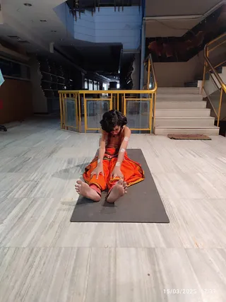 A photograph of a single person seated on a dark grey yoga mat on a pale marble floor, performing a forward-bending stretch with both hands gripping the feet. The person wears an orange-red dhoti-style garment with a golden-bordered edge and has dark, long, swept-back hair, a bare upper body, and white markings on the arms. A brown mat lies near a staircase with gold railings and a dark ceiling behind. The timestamp in the corner reads 15/03/2025 17:37.