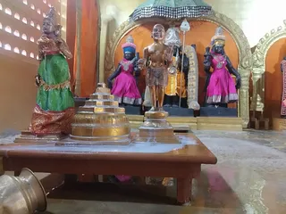 Interior shrine photo showing a wet ritual setup on a low wooden table. In the foreground a golden standing figure with raised hand, a golden pyramidal sri yantra-style meru base, and a green-and-red draped female figure stand on the table, which is coated with milky liquid as if from abhishekam. Behind them, black-stone deity murti in pink and magenta dress with garlands and crowns stand in a gilded arch, with an orange-clad figure holding a trident. A brass vessel sits at the lower left. No people are visible.