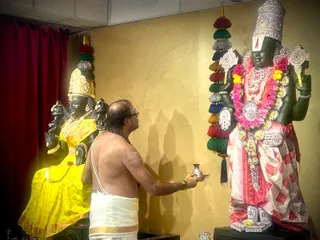 A photograph inside a temple shrine. A bare-chested priest in a white dhoti with a gold border and a brown rudraksha mala stands at the right, holding a small silver oil lamp in his right hand toward a large dark-green deity murti. The standing murti wears a tall silver crown, a red and white patterned dhoti, a pink and yellow flower garland, and gold necklaces, with a red tassel hanging beside it. To the left, another dark-skinned deity murti in a bright yellow sari and gold crown is seated against a deep red curtain. The gold-tinted wall behind them casts shadows. Two people are visible in the frame.