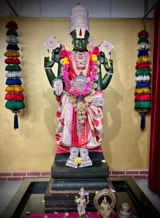 A large dark green stone murti of Venkateshwara (Perumal) stands on a black pedestal inside a temple shrine. The deity wears a tall jewelled crown with a Vaishnava urdhva pundra tilak on the forehead, a red-and-cream silk dhoti, layered gold and silver ornaments, and garlands of pink and yellow roses. Four hands hold the conch and discus. Two decorative tassel hangings in red, green, white, yellow and blue frame the shrine, and a red curtain stands at the right. Small brass and metal figures and a framed oval mirror sit on a pink cloth at the base, with scattered flower petals.