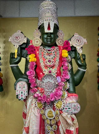 A photograph of a large dark green stone murti of Venkateshwara (Perumal) standing in a temple-style room with a gold-toned wall. The deity has four arms holding ornate silver-and-jewelled attributes, wears a tall jewelled crown with a red-and-white urdhva pundra tilak, and is adorned with layered gold and gem necklaces, a red-and-cream silk garment, and garlands of pink roses and yellow flowers. Gold and silver ornamental discs are attached across the chest and sides. No people are visible in the scene, and the statue is the sole subject.
