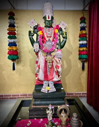 A large dark green stone deity murti of Lord Venkateshwara (Perumal) stands on a black pedestal in a temple shrine, adorned with a silver-embroidered crown with a Vaishnava tilak, a pink and pink-garlanded red and white silk dhoti and upper garment, layered garlands of pink and yellow roses, gold and silver jewellery, and silver-embroidered feet ornaments. Two colourful tassel hangings flank the murti against a gold wall, a red velvet curtain hangs to the right, and a black-edged puja tank holds rose petals. In front, a small brass utsava murti, a brass lamp and a brass kalasha sit on a pink cloth scattered with petals. No people are visible.