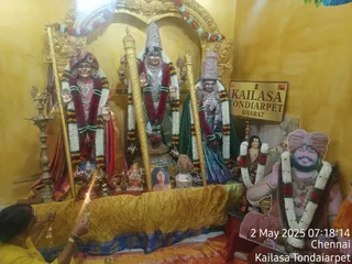 Photograph of a temple shrine with three deity murtis in an arched gold niche, adorned with white and red garlands. The central figure wears a tall silver crown and raises one hand in blessing; flanking figures wear a red-and-gold cloth and a green sari. A brass oil lamp with a flame sits at left and a brass ganesha figure with flowers and incense rests in front. A small printed photo of a woman sits on the altar. At right stands a life-size cutout of a smiling man in orange and red clothing, wearing a rudraksha mala and a white flower garland, with a gold-painted sign reading KAILASA TONDIARPET BHARAT on the wall. A woman in yellow lights a lamp in the lower left corner. Timestamp shows 2 May 2025, Chennai, Kailasa Tondaiarpet.