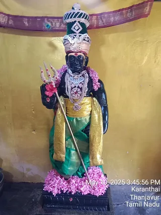 A photograph of a standing black stone deity murti in a temple shrine, shown frontally against a golden-yellow wall with a maroon and gold patterned cloth border above. The murti wears a tall green and silver ornate crown, a silver and jewelled necklace with red stones, a green silk dhoti with a wide gold border and gold sash, and holds a silver trident adorned with red stones and a red flower. Pink flower garlands drape the shoulders and the base, which rests on a dark stone pedestal with red dots. A timestamp and location overlay reads May 10, 2025, Karanthai, Thanjavur, Tamil Nadu.