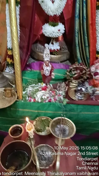 A photograph of a shrine altar decorated with white jasmine garlands and red-and-yellow flower strands, with a small metal deity murti dressed in a red garment and silver crown and necklaces standing on a green silk cloth. Scattered white and red flowers and green leaves lie in front of the figure. On the foreground table a lit oil lamp, a brass kalasha-style vessel with water, small clay cups and a bowl of green leaves sit beside it. A brass bell and a lamp stand are at right. No people are visible. A timestamp and address overlay reads 10 May 2025 at 42A Kasima Nagar 2nd Street, Tondiarpet, Chennai, Tamil Nadu.