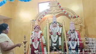 A photograph inside a temple shrine with a golden arched niche decorated with flower garlands in red, white and blue. Three deity murtis stand in the niche: a figure with a red turban and white garland on the left, a central figure in a silver crown with a green backdrop, and a figure in a silver crown and red-and-green sari on the right. A woman in a pale yellow top stands at the left edge holding a brass oil lamp (deepa) toward the murtis, as in an aarti. Garlands drape the central figures and the walls are yellow.