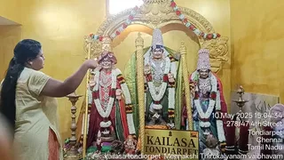 A woman with long dark hair in a pale yellow kurta stands at left, extending her right arm toward a shrine. Three deity murtis are decorated with white and red flower garlands in a gilded arched niche. The central murti wears a tall conical crown and a green silk backdrop; the left and right murtis wear crowns, gold ornaments and red and green garments. Brass oil lamps stand on either side, and a golden placard reads KAILASA TONDIARPET. The wall is painted yellow and a timestamp reads 10 May 2025.