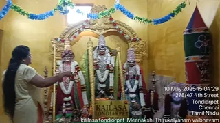 A woman in a cream and gold sari stands at left, long dark hair falling down her back, reaching toward a richly decorated shrine. Three figures are seated on a golden platform in a yellow-walled room: the central figure wears a tall conical crown and a green garment with a white flower garland, while the figures on either side wear red and gold garments, tall crowns and garlands of white and orange flowers. A gold plaque at the base reads KAILASA TONDIARPET. Blue and green paper streamers hang across the top, and a gold arch frames the shrine. At right stands a tall red, green and blue banner with decorative symbols, and a small figure in pink sits near the bottom right. A date and address stamp appears in the lower right corner.
