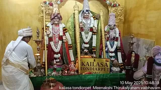 A worship scene before three large statues on a green-draped altar in a gold-walled shrine. On the left, a standing figure in a red cloth and white garland holds a raised hand. In the centre, a figure in a white, gold and green garment with a tall crown and white garlands holds a golden staff. On the right, a figure in a tall decorated crown and red garland stands beside a gold staff. In the foreground at left, a person in white ritual dress with a white head wrap bends over the altar, tending oil lamps and brass vessels. A small Ganesha murti sits on the altar, and a painted figure at the far right wears a rudraksha-style garland. A gold plaque reads KAILASA TONDIARPET, and a timestamp shows 10 May 2025.