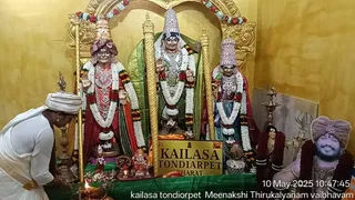 A photograph inside a shrine with a gold-toned backdrop showing three silver-finished deity murtis draped in green cloth and white and red garlands. The central figure holds a raised hand and a tall crowned headdress; the figures on either side wear crowns and jewelled necklaces. A gold sign reads KAILASA TONDIARPET BHARAT. In the lower left a man in white ritual dhoti and white head cloth bends forward over offerings, with a brass oil lamp burning at the front. On the right a framed portrait shows a smiling man with a saffron turban, rudraksha-style beads and garlands, labelled with a date-stamped caption at the bottom. Brass lamps, a kalasha-like vessel and ganesha figure sit among flowers at the base.