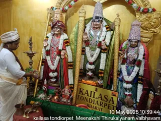 A photograph of a temple shrine with three decorated murtis (deity statues) draped in white and orange-red garlands, set before a gold-framed backdrop. The central figure wears a silver crown and green silk, the left figure a red and gold robe with a turban-like headdress, and the right figure a purple sari with a silver crown. A gold-lettered board in front reads KAILASA TONDIARPET BHARAT. A woman in a white dhoti and white head covering stands at the left edge, holding a lamp and tending offerings. Brass oil lamps, a small Ganesha figure, flower petals and a kalasha sit on a green cloth. A framed photograph of a person in saffron appears at the lower right corner, but it is a printed picture and not a physical person.