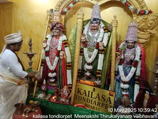 A photograph inside a temple shrine. A woman in a white dhoti-style garment and white head cloth stands at left, bending over a tray of flowers beside brass oil lamps. Three large deity murtis stand on a green cloth in a golden-walled niche, each garlanded with white and orange-red flower strands. The central figure is adorned with ornate jewelry and a tall crown, the left figure wears a red and gold garment with a tall gold crown, and the right figure wears a blue-green sari and a crown. Golden staffs stand beside the murtis, and a small Ganesha figure, a kalasha and a brass pot sit at the base. A golden placard reads KAILASA TONDIARPET BHARAT. Timestamp text at bottom right reads 10 May 2025 10:39:42.