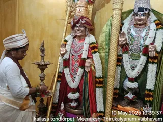 A photograph inside a temple shrine. On the left, a woman in a white dress with an orange-bordered sari and a white head wrap stands before a tall brass oil lamp (deepam) and holds a small plate of flowers and offerings, which she is lowering toward the lamp. Behind her stand two large decorated deity murtis with golden raised hands. The left murti wears a red and gold dress, a red and gold crown, a red-and-pink jewelled necklace and a long garland of white jasmine and orange-and-red flowers. The right murti wears a green garment, a silver crown, a silver jewelled necklace and a similar white and orange garland. Golden pillars frame the shrine, and a bottom-right caption reads 10 May 2025 10:39:37 Meenakshi Thirukalyanam vaibhavam.