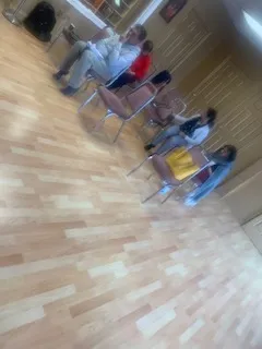A blurry, tilted indoor photograph of a room with light wooden laminate flooring. Several people sit on metal folding chairs in a loose group. One person in the centre wears a red top and another wears a yellow garment, while figures at the edges are partly cropped. Wood-panelled walls and a doorway are visible in the background.