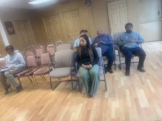 A photograph inside a room with wooden flooring and cream walls, where several people sit in rows of folding chairs with pinkish-beige upholstery. In the foreground a woman in a dark top and teal-green trousers sits upright with her hands together. Beside her a man in a blue shirt sits with his hands clasped, and further right another man in a light blue shirt sits on a chair. On the far left a man in a dark jacket sits near an empty row of chairs. Doors and a small framed picture are visible on the back wall.