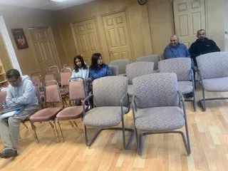 A photograph of a small room with light wooden laminate flooring and tan walls with wooden doors. Rows of grey upholstered chairs and pink-cushioned folding chairs are set out in a loose arrangement. Several seated people are visible: a man in a grey shirt at the lower left, two women in blue and dark tops sitting together near the middle, and two men in blue and dark shirts seated at the upper right. A small framed picture hangs on the wall at the left edge.