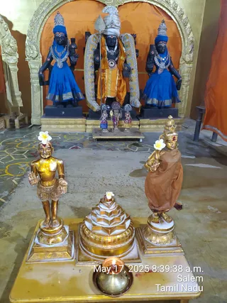 Photo of a temple shrine with dark stone deity murtis in an arched golden niche backed by orange drapery. The central standing murti wears an orange dhoti, white turban, silver-and-jewel ornaments and holds a trident. Two flanking murtis wear blue skirts, tall silver crowns and garlands. In front on a marble platform stand two gold-painted figures, each with a white flower, and a white-and-gold sri yantra with a flower on top. A small brass oil lamp burns at the front. The floor has yellow and white floral painted patterns. A timestamp reads May 17, 2025 8:39:38 a.m. Salem, Tamil Nadu.