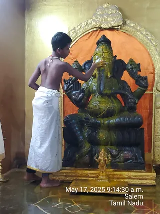 A bare-chested young boy in a white dhoti with a white sacred thread across his shoulder stands at the left, placing his right hand on the forehead of a large dark green-black stone murti of Ganesha. The seated Ganesha has an elephant trunk, a crown, and four arms, and sits on a carved pedestal before an orange cloth backdrop framed by a gold arch. A small gold Ganesha figure is set on the base in front. The floor is wet and has a faint kolam design. A timestamp reads May 17, 2025 9:14:30 a.m., Salem, Tamil Nadu.
