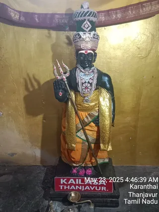 A standing black stone deity murti decorated for worship, shown against a gold-painted wall with a maroon-and-gold cloth canopy at the top. The figure wears a tall ornate crown studded with silver and red stones, a white-and-red forehead mark, a silver necklace with a large central pendant, and a gold-bordered orange dhoti with a gold shawl. It holds a silver trident with red-dotted prongs in one hand. The deity stands on a dark pedestal with pink lotus-like feet, and a brass oil lamp sits in front. A printed red sign on the base reads KAILASA THANJAVUR. A timestamp reads May 23, 2025 4:46:39 AM with Karanthai, Thanjavur, Tamil Nadu.