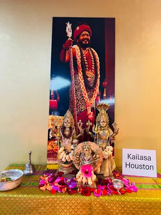 A photograph of a shrine table with a tall printed portrait on the wall of a bearded man in a red turban, leopard-print garment, orange-and-white flower garland and rudraksha mala, holding a silver trident. a person appears only as this printed portrait, not a physical person. Before it stand brass deity murtis, including a central Kalabhairava-style figure with a dog and a flanking pair of figures with tridents, surrounded by scattered pink, red and purple flower petals. A silver bell, a brass bowl and a small silver vessel sit on a gold-and-green brocade cloth. A white card reads Kailasa Houston. No physical people are visible.