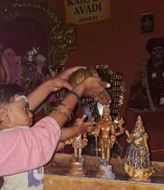 A child with white vertical forehead markings in a pink top stands at a shrine, reaching toward small golden deity murtis on a raised ledge. An adult woman with bangles pours or lifts a brass vessel above the figures, with water visible. Three gilded figures are shown: a smaller one on the left, a central four-armed figure with a crown, and a figure in a blue and gold sari on the right. Behind them is a gilded arch and a carved throne, with pink and white flower garlands. A sign on the wall reads KAILASA AVADI and BHARAT. A framed picture of a smiling man in dark clothing with a garland and a rudraksha mala appears at the upper right, but he is in a portrait, not physically present.