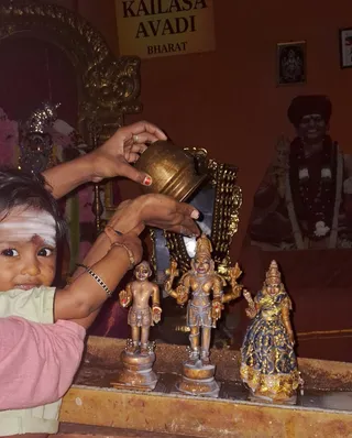 A photograph inside a temple shrine room at KAILASA Avadi, Chennai. A small child with a white sacred ash band across the forehead and a red mark between the brows, wearing a pink top and pale green shirt, looks toward the camera at the left edge. An adult's hand with red nail polish and a ring holds a brass pot pouring liquid, while another hand touches the bronze and gilt deity murti set on a stone ledge: a small standing figure, a four-armed standing deity murti wearing a crown and holding objects, and a female figure in a blue and gold sari. A framed portrait on the right wall shows a man in a dark turban garlanded with white flowers and beaded necklaces. A yellow signboard above reads KAILASA AVADI BHARAT. Orange walls and gilded ornate frames surround the scene, with a pink floral decoration at the left.