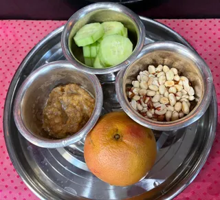 Overhead photo of a round stainless steel thali tray on a pink polka-dot cloth. Three small steel bowls hold sliced cucumber, a brownish mashed paste, and peeled groundnuts with reddish skins. A whole orange-red citrus fruit sits at the centre of the tray. No people or deity visible.