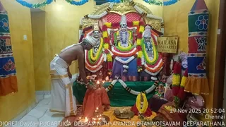 A photograph inside a yellow-walled shrine room. A shirtless priest in white dhoti with gold border and a white head cloth bends forward at the lower left, lighting a row of oil lamps on a brass plate in front of a red-and-gold altar. Three deity murtis stand on a green-draped platform, decorated with white, yellow and orange garlands; the central figure has a silver crown and is flanked by two others. A brass tray with offerings sits on an orange cloth at right, and a small signboard reads KAILASA THONDIARPET BHARAT. A large framed portrait of a bearded figure in a saffron-coloured turban appears at the right edge, partly obscured by garlands and a decorative red and green banner. A timestamp reads 29 Nov 2025 20:52:04 Chennai, and a lower banner reads Day6 Deepam Nithyananda Brahmotsavam Deeparathana.