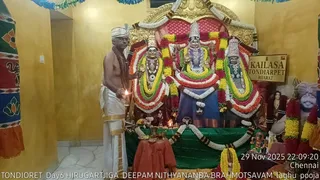 A photograph of a ritual in a small shrine room with yellow walls and a marble floor. A bare-chested priest in a white dhoti and white head cloth stands at left holding a lit oil lamp in his right hand, with a rudraksha-style cord across his chest. Behind him is an ornate golden shrine with three deity murtis adorned with garlands of orange, yellow and white flowers; the central figure wears a tall crown. A signboard reads KAILASA TONDIARPET BHARAT. A green cloth covers the altar in front, with brass vessels, a kalasha, lamps and a flower garland. A garlanded portrait is visible at right. The timestamp reads 29 Nov 2025 22:09:20 Chennai, and a caption reads TONDIORET Day6 HIRUGARTJIGA DEEPAM NITHYANANDA BRAHMOTSAVAM laghu pooja.