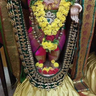 A close-up photograph of a deity murti adorned for worship. The figure wears a magenta silk garment with gold border, a green and gold embroidered shawl draped over the shoulders, and a large crown-like silver ornament set with a green stone at the chest. Layers of yellow marigold garlands with red rose accents hang from the neck and waist, along with silver bead necklaces. Yellow and red flowers are arranged in front of the base, with a silver hand visible at upper right. Yellow silk fabric lies at the bottom. No people are visible.