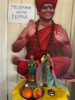 A home shrine with a large life-size cutout of a smiling man in a red turban and red robes, seated cross-legged with both palms raised in blessing, wearing long rudraksha malas and red rudraksha bracelets. A handwritten paper sign reads 'MELBOURNE HOME TEMPLE'. In front, on a yellow cloth, stand two small gilded murti figures: one in a teal outfit with a white-beaded red turban, the other in a green embroidered sari with a veil and a red rose, with a green parrot perched beside it. A steel bowl holds an orange and an apple, and a small oil lamp with a lit wick sits to the right.