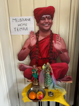 A photograph of a home altar set up in front of a cream-painted door. Behind the altar stands a large cutout figure of a smiling man seated cross-legged, wearing a red turban, red robes, a dark red rudraksha mala with a gold necklace, and red beaded bracelets; his palms are raised in blessing. A handwritten paper sign pinned to the door reads MELBOURNE HOME TEMPLE. In front, a small brass deity pair stands on a pedestal: one in a teal garment and one in a green embroidered sari with a sheer veil and a green parrot perched on the figure's shoulder, with a red flower at the waist. Before them sit a stainless steel bowl holding an orange and an apple, a small silver oil lamp, and a yellow cloth spread across the base. No living people are visible.