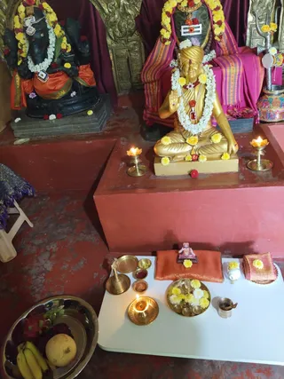 Overhead view of a shrine with no visible living people. Centre: a gilded statue of a seated figure in a golden turban with a white flower garland and yellow marigolds, raising one hand in blessing, with a red ornament on the chest. Behind it, a magenta cloth backdrop and striped textiles frame the statue. To the left, a dark stone murti of an elephant-headed deity wears a white and red garland and an orange cloth. On the white ritual table in front: a brass bell, small brass bowls, a folded orange cloth, a brass plate of marigolds and flowers, a small brass lamp with a lit oil flame, and a metal cup. Two brass oil lamps burn beside the statue, and a brass bowl of fruit including bananas and dragon fruit sits at lower left.