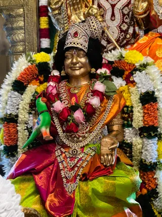 A photograph of a golden-toned statue of a seated female deity, adorned with a jeweled crown set with a red stone, ornate gold and gemstone necklaces, and a bright orange and magenta silk sari with a lime-green lower garment. The statue is garlanded with red and pink roses and green leaves, with a green toy parrot perched at its left side. Behind it are marigold and white flower garlands in orange, yellow and white, along with gold-embroidered red and orange fabric. The figure's right hand rests open on its lap, and its eyes are closed in a serene expression. This appears to be a decorated murti in a worship setting, consistent with an alankar display.