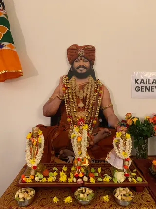 A life-size cutout of a seated man in a rust-orange turban with a gold ornament, wearing orange robes, layered gold and pearl necklaces, rudraksha beads and gold armlets, sits in padmasana with hands in a blessing gesture against a white wall. Flanking him are two small gold-toned murti figures, each draped in white and yellow flower garlands, with a central murti also garlanded. Yellow and white flowers are scattered on a red and gold altar cloth, with three steel bowls of cut fruit in front and a vase of orange and yellow roses at right. A partial sign reading KAILA... GENEV... is visible at right, and orange and green cloth hangs at upper left. No physical people are present.