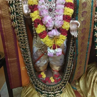 A photograph of a deity murti in a temple shrine, shown from the waist down with a flower garland of pink lotus blossoms, red roses and yellow marigolds draped over its upper body. The figure wears a silver-white dhoti with gold brocade borders and gold ornaments, and its raised silver hands are visible at the sides. Fresh red roses and yellow marigolds are scattered at its base, and the murti is framed by a green and gold embroidered cloth with a red and gold patterned border. No people are visible.