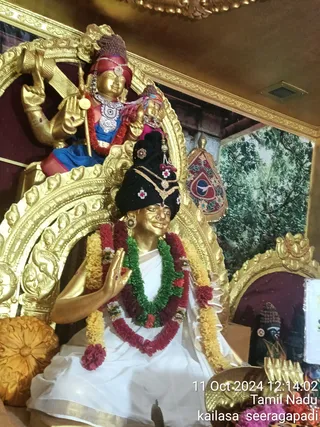 A gilded statue of a seated figure in a white dhoti-style garment with a black turban adorned with gold jewellery and small red-and-white flowers, wearing layered garlands of yellow, red and green flowers, its right hand raised in blessing, seated within an ornate gold arched frame. Behind and above, a gilded deity murti in a blue garment with a red headcloth, holding a trident-like staff and a silver-toned necklace, with a smaller figure beside it. Right background shows a painted forest scene, a red and gold ceremonial pennant, and a small dark murti. Photo timestamp overlay reads 11 Oct 2024 and Tamil Nadu.