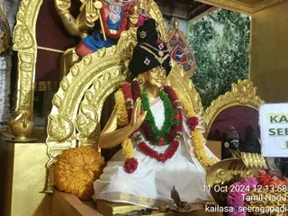 A golden statue of a seated figure in a gilded shrine is the main subject. The figure wears a tall black turban decorated with a jewelled gold ornament, a white dhoti with a gold border, and layered garlands of yellow, red and green flowers. The right hand is raised in a blessing gesture. Behind it stands a golden deity murti in blue and red drapery with a trident, and a painted forest backdrop fills the right side. Brass oil lamps, a golden cushion and small offering bowls sit on the shrine platform. The statue is a depiction, not a living person, so no one is physically present.