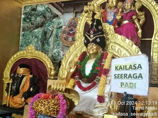 A photograph of a golden deity murti seated on a throne in a temple shrine. The figure wears a white dhoti with a black and gold headdress decorated with red flowers and a gold ornament, and is draped in green, red and yellow garlands. Its raised right hand is in abhaya mudra. Beside the throne sits a black stone murti of a seated figure with an elephant-like head, wrapped in an orange cloth. Two gold deity figures with crowns appear above in a golden arch, one in blue and one in pink. A leopard-print cloth lies in front with a garland and fruit offerings. A white sign reads KAILASA SEERAGA PADI. A camera timestamp reads 11 Oct 2024 and Tamil Nadu. No people are visible.