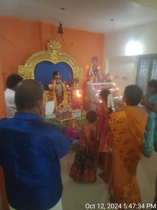 A photograph inside a shrine room with orange walls and a gold-framed arch holding a blue backdrop, where a deity murti stands dressed with garlands and a yellow shawl. A lamp flame burns before it. To the right, a white-draped altar stage displays small painted figures and a portrait with a garland on top. A sign reading KAILASA AVADI sits at the base of the altar. Several devotees stand and kneel in puja, including a man in a blue shirt in the foreground, a woman in an orange sari with hands joined, and a woman in red holding a flame or tray. Another woman in teal stands at the right edge. A timestamp in the corner reads Oct 12, 2024 5:47:34 PM.