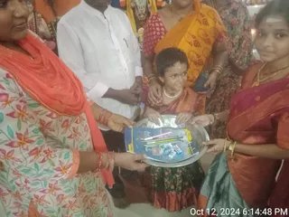 A photograph of a group of women and a young girl gathered around a silver tray held by two women. The tray holds pens, pencils, a small blue-and-yellow notebook or pack and a few small boxes. The woman on the left wears a white and orange floral salwar with an orange dupatta and many bangles. A young girl in the centre wears a red and green half-saree, a pearl necklace and a bindi. A man in a white shirt stands behind them, and a woman in a yellow-and-orange saree with bangles stands at the upper right. A woman in a pink-and-red saree holds the right side of the tray. The background shows bright decorations and a timestamp reading Oct 12, 2024 6:14:48 PM.