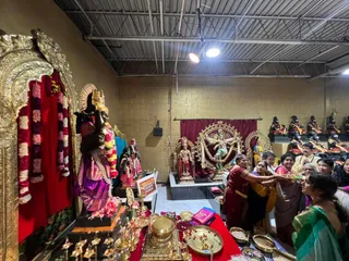 Photograph inside a hall with a corrugated metal ceiling and tan block walls, showing a temple shrine lined with gold-framed arches and red-and-pink garlands. A large dark deity murti in a crown and purple-and-gold dress stands at left, with a dancing Nataraja-style bronze and a smaller deity murti in pink at centre on a platform. A sign on a stand reads KAILASA TORONTO CANADA. In the foreground a brass plate of offerings and small oil lamps sit on a red cloth. Several women in saris, one in a maroon sari and others in yellow, pink and green, stand close together and reach toward the offering tray. Rows of seated figures are visible in the background at right.