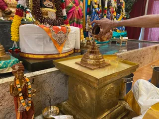 Temple abhishekam scene: a hand pours water from a copper kalasha over a small copper shrine or lingam-like form set on a large brass pedestal with a golden top. Behind it is a decorated altar with a deity murti draped in saffron and white cloth, adorned with gold ornaments, rudraksha beads and marigold garlands. A brass lamp with incense sticks sits in front, and a brass figure of a standing attendant wearing an orange sari and marigold garland stands at left. Pink and green silk saris and blue fabric drape the background shrine.