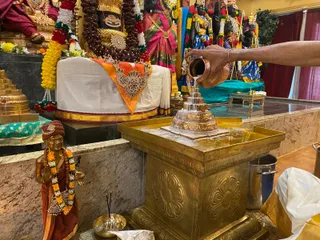A photo inside a temple shrine showing a priest's arm, bare except for a bangle, pouring water from a copper pot over a silver shivalingam set on a large brass base. Behind the lingam stands a white cloth draped over a pedestal with an orange and gold ornamented sash, and to the left a brass pillar stands with gold and red tiers. Gold and rudraksha-bead garlands hang on the deity murti behind. A small brass figure of a standing devotee in saffron-red dress with a flower garland stands to the left, and a brass oil lamp and incense sticks sit on the base. Marble-topped altar surfaces, colourful pink, green and gold textiles and additional deity murtis with crowns and garlands fill the background. This shows abhishekam being performed.
