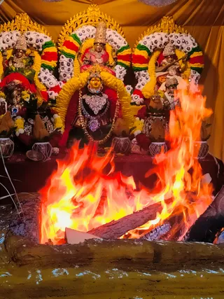 A large fire blazes in a clay-rimmed homa pit in front of a decorated altar with several deity murtis. The central murti wears a silver-embellished red garment, a jeweled crown and layered silver necklaces, seated under a golden arch wrapped in yellow, red, green and white flower garlands. Flanking murtis wear silver headdresses and crowns, and small brass vessels with mango leaves and coconuts stand on the altar. Gold drapery forms the backdrop. No people are visible.