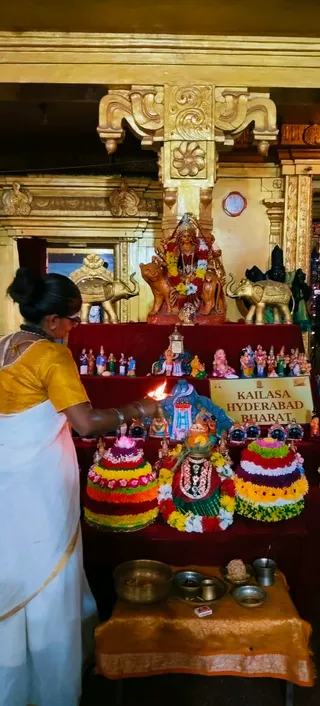 A woman in a white sari with a mustard-yellow blouse lights an oil lamp at a decorated shrine inside a gilded temple hall. She has her hair in a bun and wears glasses. Behind her stands a golden-brown deity murti seated on a lion-like mount, garlanded with marigolds, flanked by two gold-and-silver elephant figures. Tiered cones of red, yellow and white flowers stand on a maroon cloth, with a green-and-red garlanded idol in the centre. Small clay dolls, a brass bowl and plates with offerings sit on a gold-bordered table below. A sign reads KAILASA HYDERABAD BHARAT. The scene suggests a Navaratri worship setting.