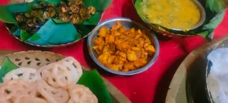 A close-up photo of a festive food spread on a red cloth. In the upper left, fried okra-like pieces sit on a green banana leaf inside a metal plate. In the center, a small steel bowl holds cubes of spiced golden-brown fried potato or paneer-like pieces. At the upper right, a bowl of yellow dal or curry rests on a leaf. At the lower left, white jalebi-like lacy rings lie on a banana leaf over a metal tray, and at the lower right a plate holds white rice-based items. No people are visible.