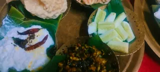 A close overhead photo of a traditional vegetarian meal served on a brass plate with banana leaf liners. On the left is a mound of white curd topped with two dried red chillies and tempering. Upper right shows sliced cucumber in a perforated brass bowl. Bottom centre holds a dry vegetable stir-fry with green and brown pieces in a brass bowl. Crisp white wafers sit at the top edge on a banana leaf. A red cloth is visible at the lower right. No people are visible.